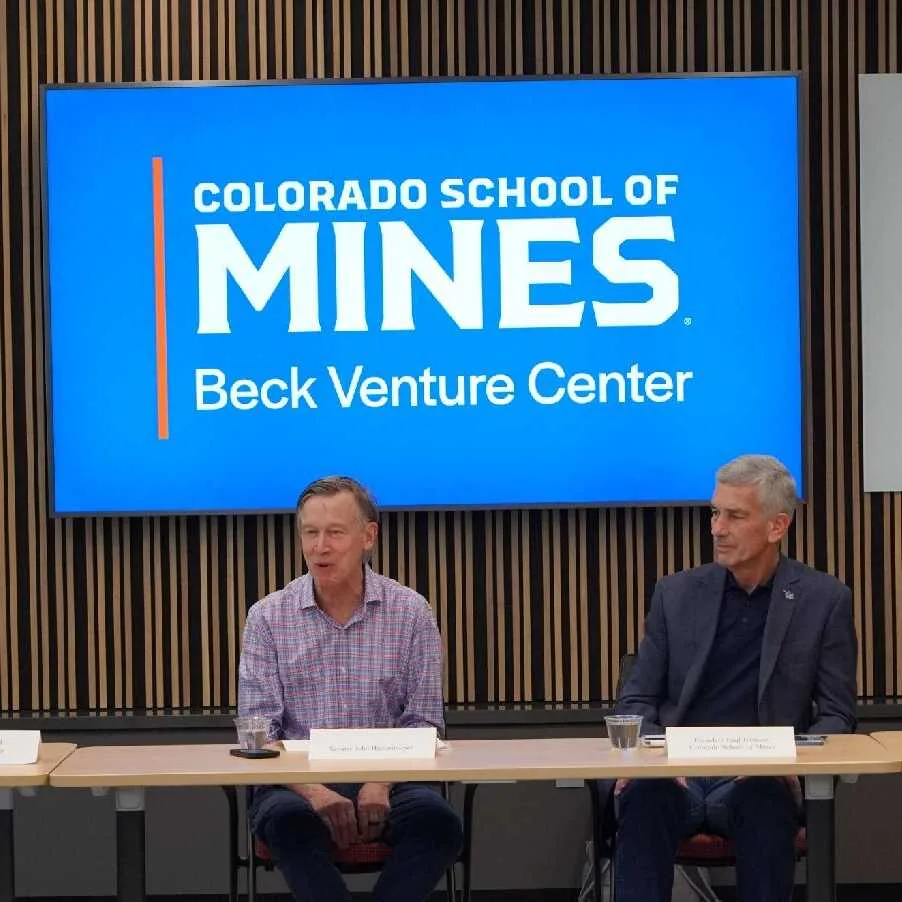 Sen. Hickenlooper and Mines President Paul Johnson seated at a table in front of a Colorado School of Mines Beck Venture Center sign during a panel discussion.