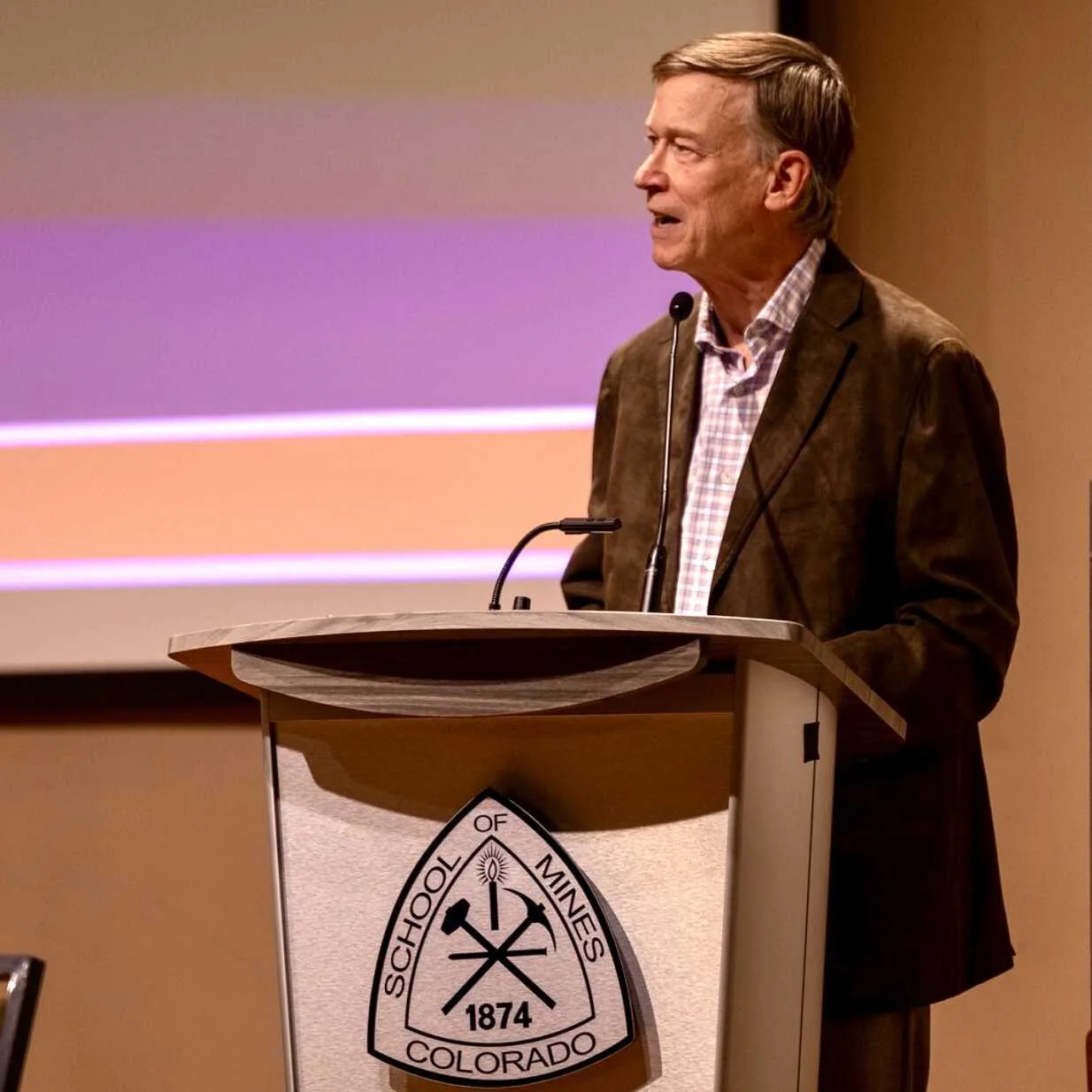 Sen Hickenlooper in a brown jacket speaking at a podium with the Colorado School of Mines logo during a presentation.