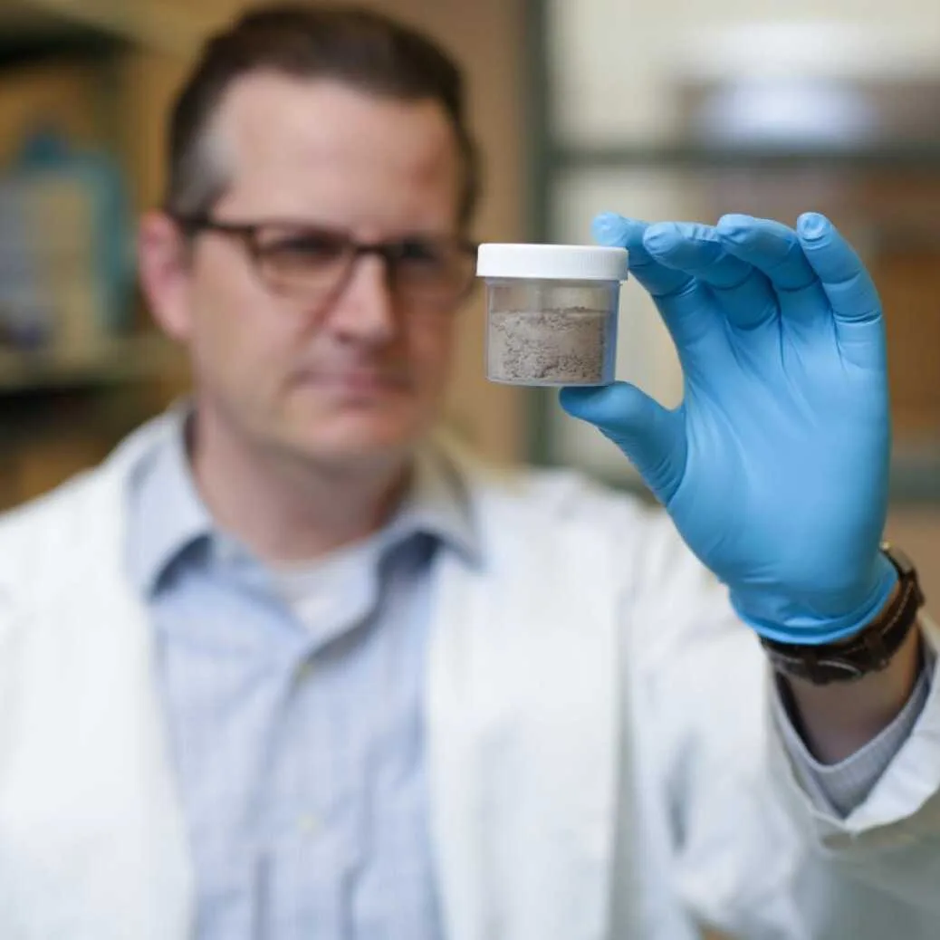 Chris Higgins in lab coat and blue gloves holding a small container with a gray powder substance up for examination.