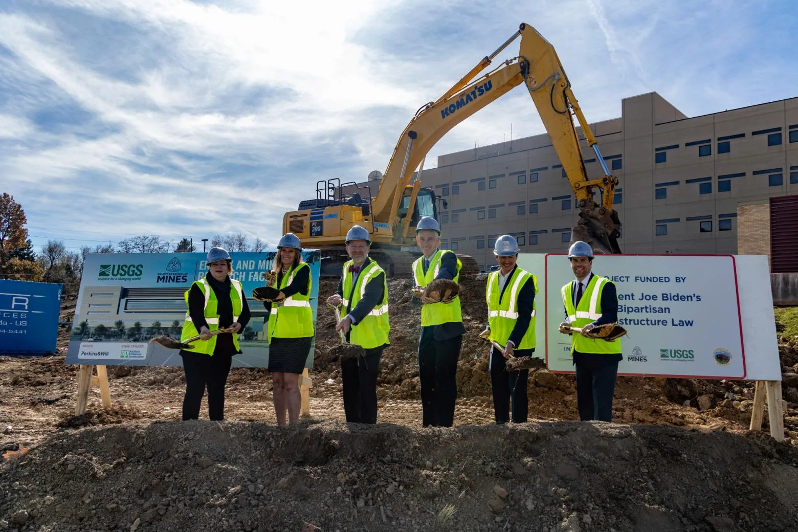 Six people in safety vests and helmets hold shovels during a groundbreaking ceremony at the USGS building construction site with an excavator and signage.