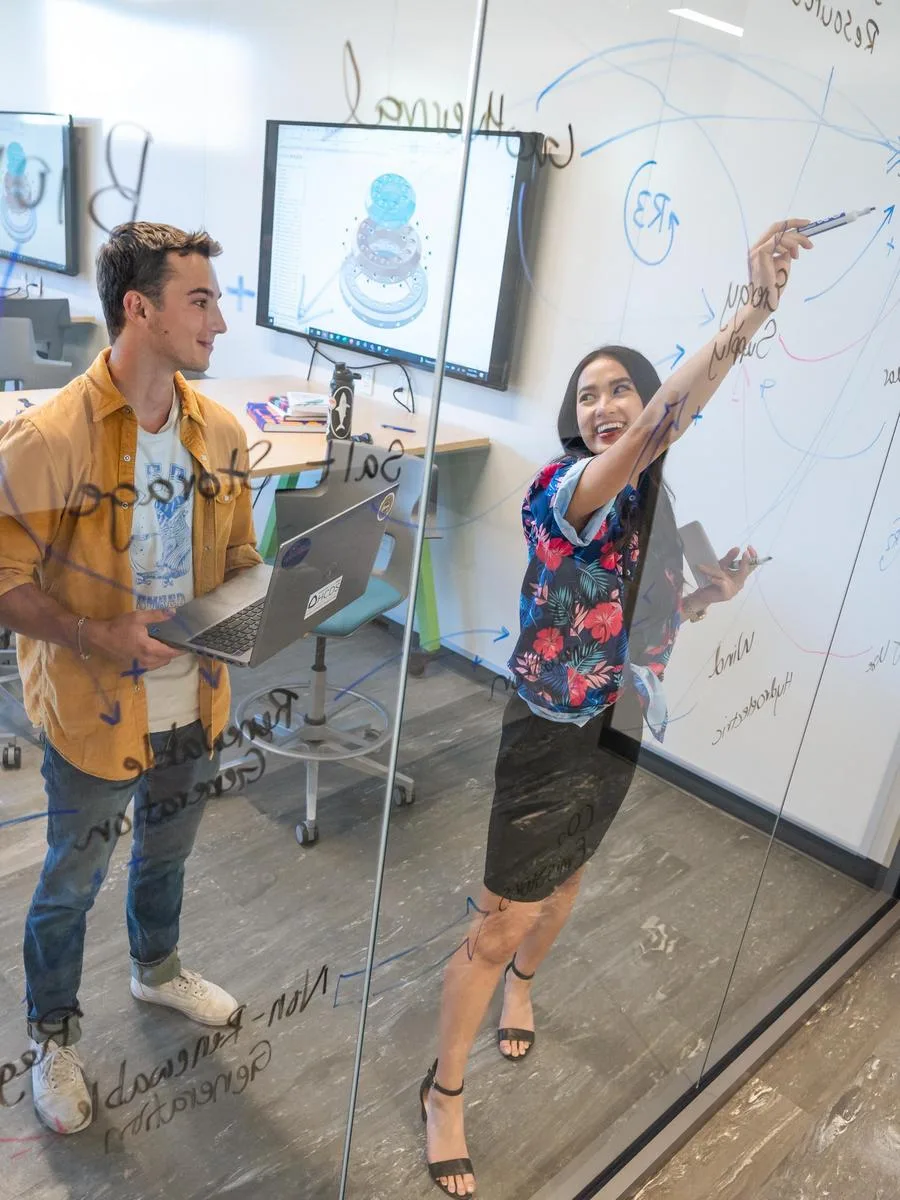 A senior Mines student draws equations on a clear wall in a conference room explaining her concepts to another student