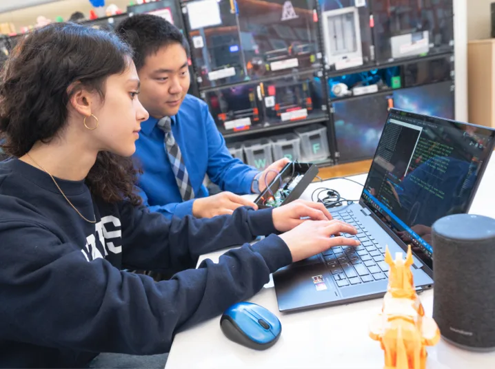 This image shows students collaborating on a technical project, with one person typing on a laptop while the other observes.