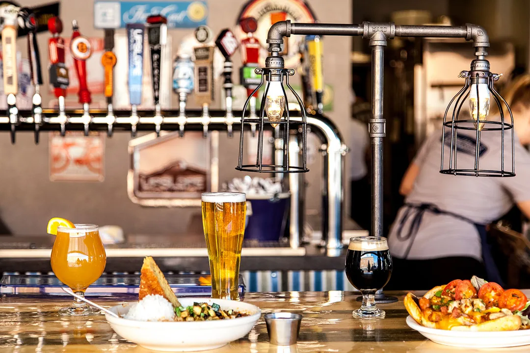Bar counter with three different beers and plates of food, with beer taps and industrial-style hanging lights in the background.