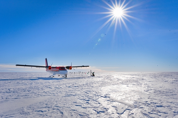 Research plane sits on Antarctic ice with bright blue sky and sun in background