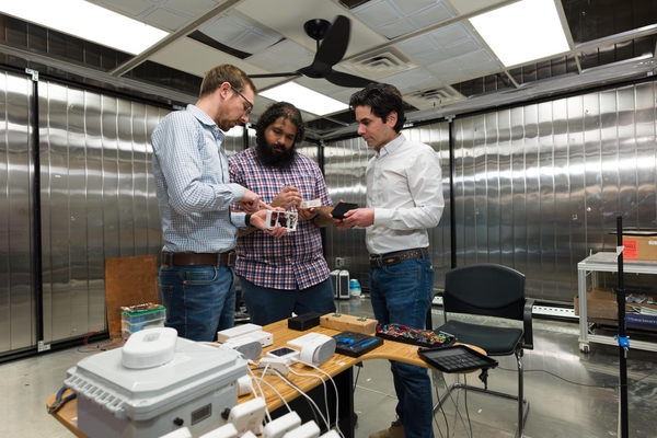Three researchers in a metal-paneled engineering lab gather around a wooden worktable, inspecting small electronic devices and circuit prototypes.