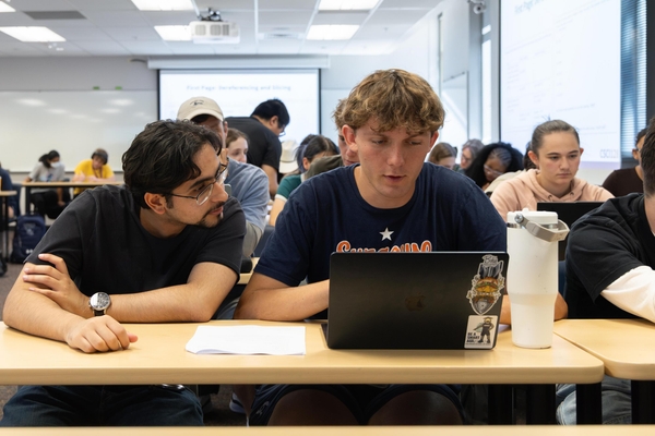 Two students look at a laptop screen on the desk in front of them during class