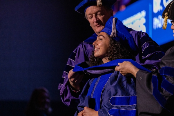 A university official in blue and purple regalia hoods a smiling graduate on stage during a Colorado School of Mines commencement ceremony.