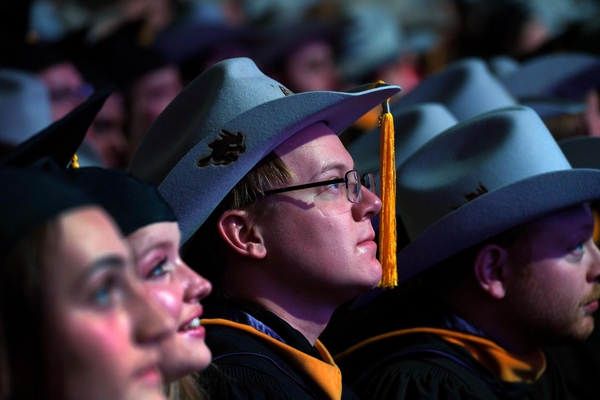 Mines graduate student wearing a felt Orediggers cowboy hat with gold tassel listens during commencement, surrounded by fellow graduates in academic regalia.