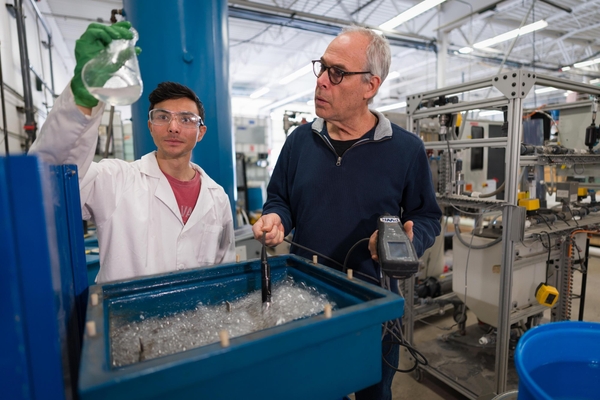 A graduate student wearing safety glasses holds up a flask of clear liquid while an older professor dips a probe into a tub of aerated, bubbling water in a testing lab.