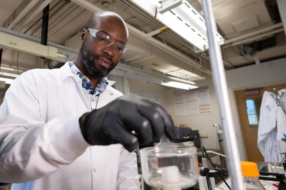 A student in a white lab coat and safety glasses works in a lab