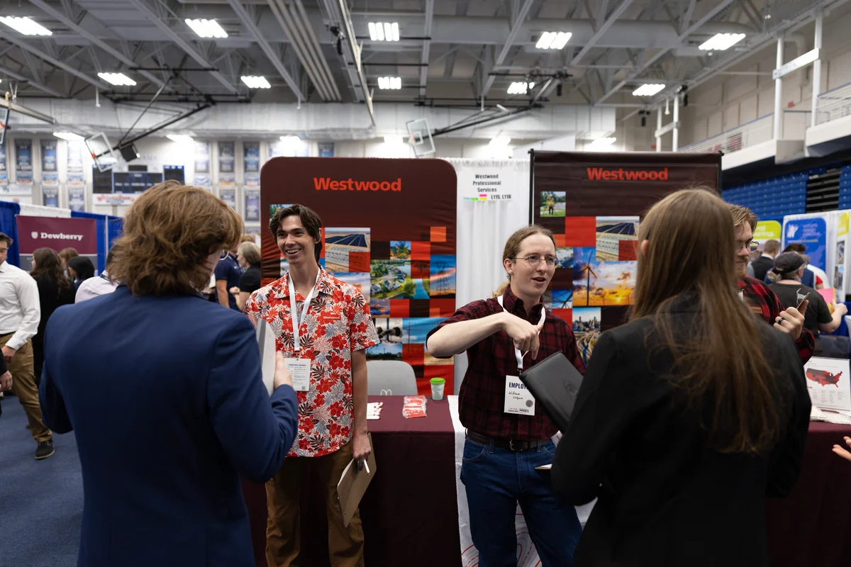 Two students in business attire talk with two Westwood Professional Services recruiters, one in a red floral shirt, at a booth in a crowded gym