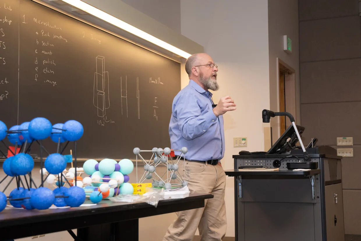Mines faculty member Gerald Bourne giving a lecture in a classroom with molecular models on a table and diagrams drawn on a chalkboard behind him.