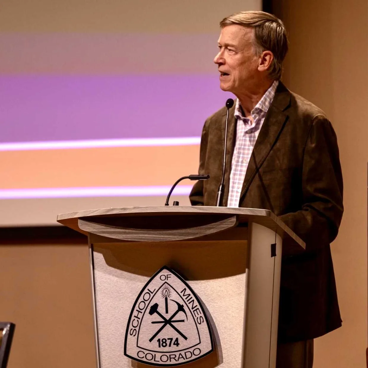 Sen Hickenlooper in a brown jacket speaking at a podium with the Colorado School of Mines logo during a presentation.