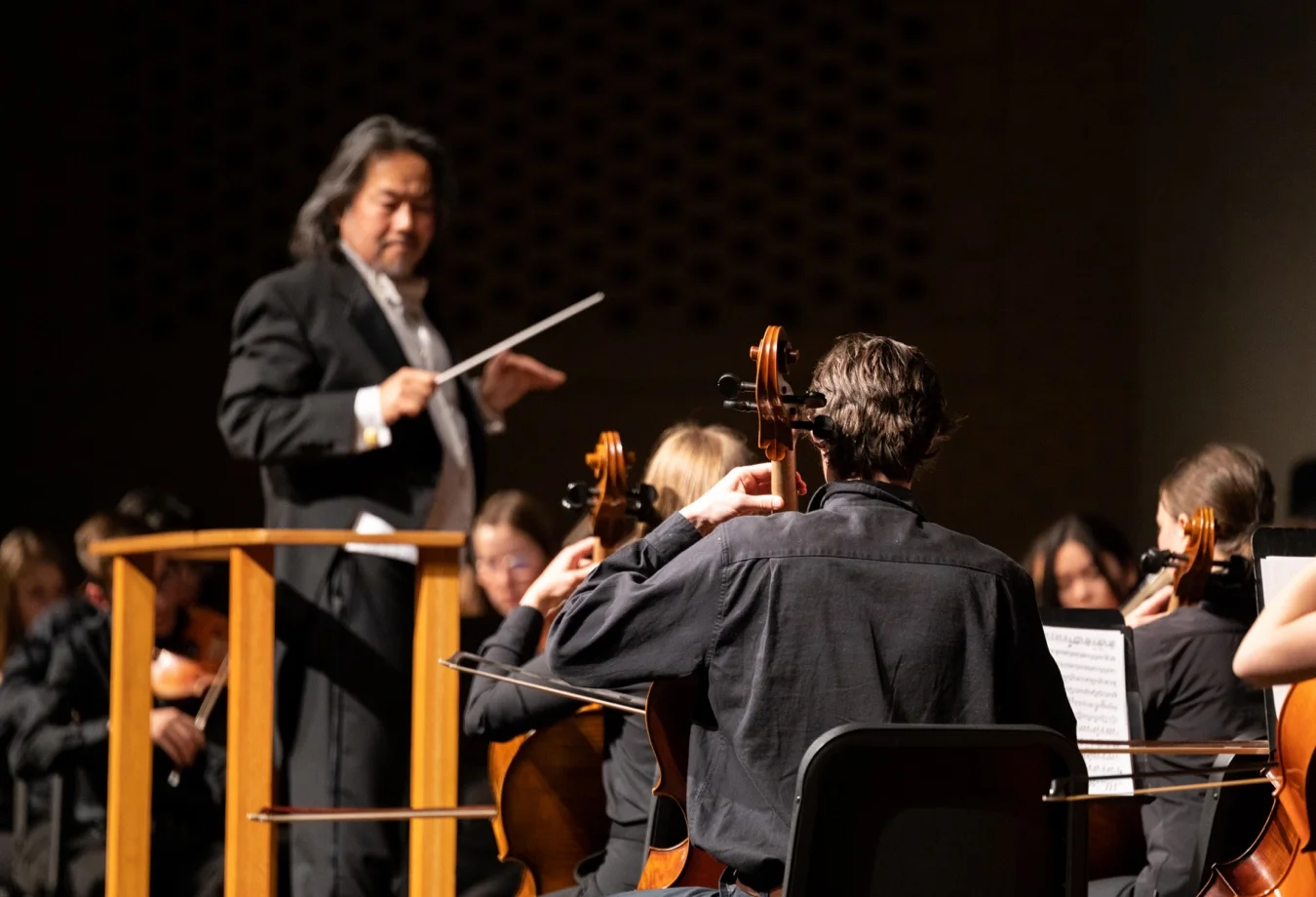 This image shows a symphony conductor in a black suit directing an orchestra from behind a wooden music stand.