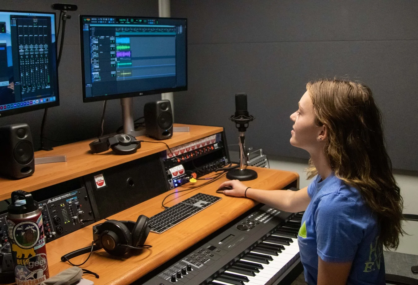 A female Mines student working in a recording studio featuring a wooden desk setup with multiple monitors, speakers and an MIDI keyboard controller.