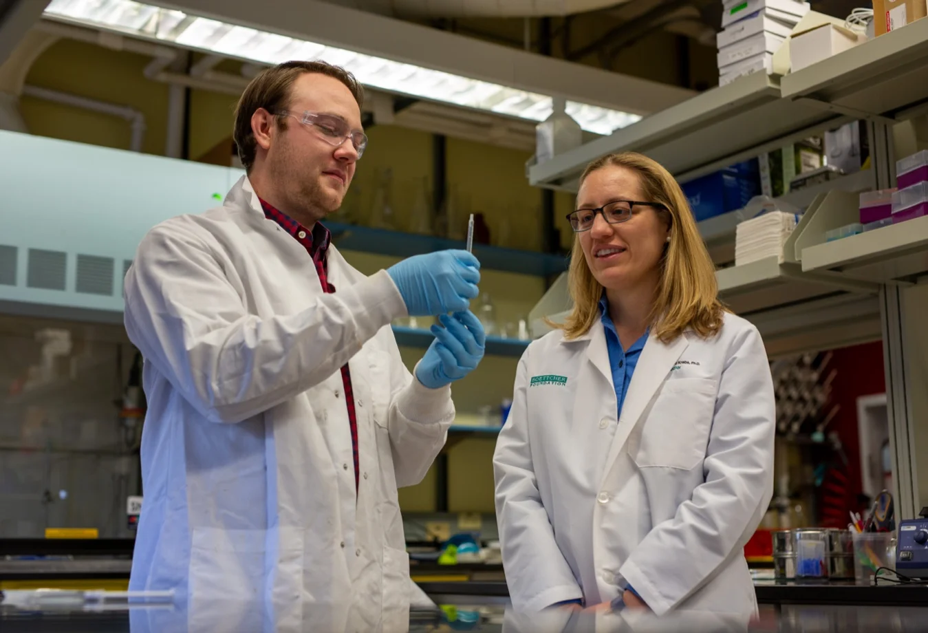 A male student with a white lab coat on performing a test, while the professor in a white lab coat watches in a lab setting.