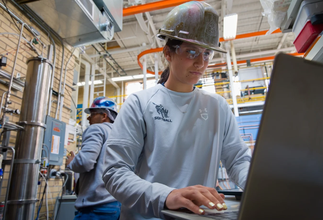 A female student, in an industrial workspace, with a hard hat on working on a computer with a male student behind her reading test results.