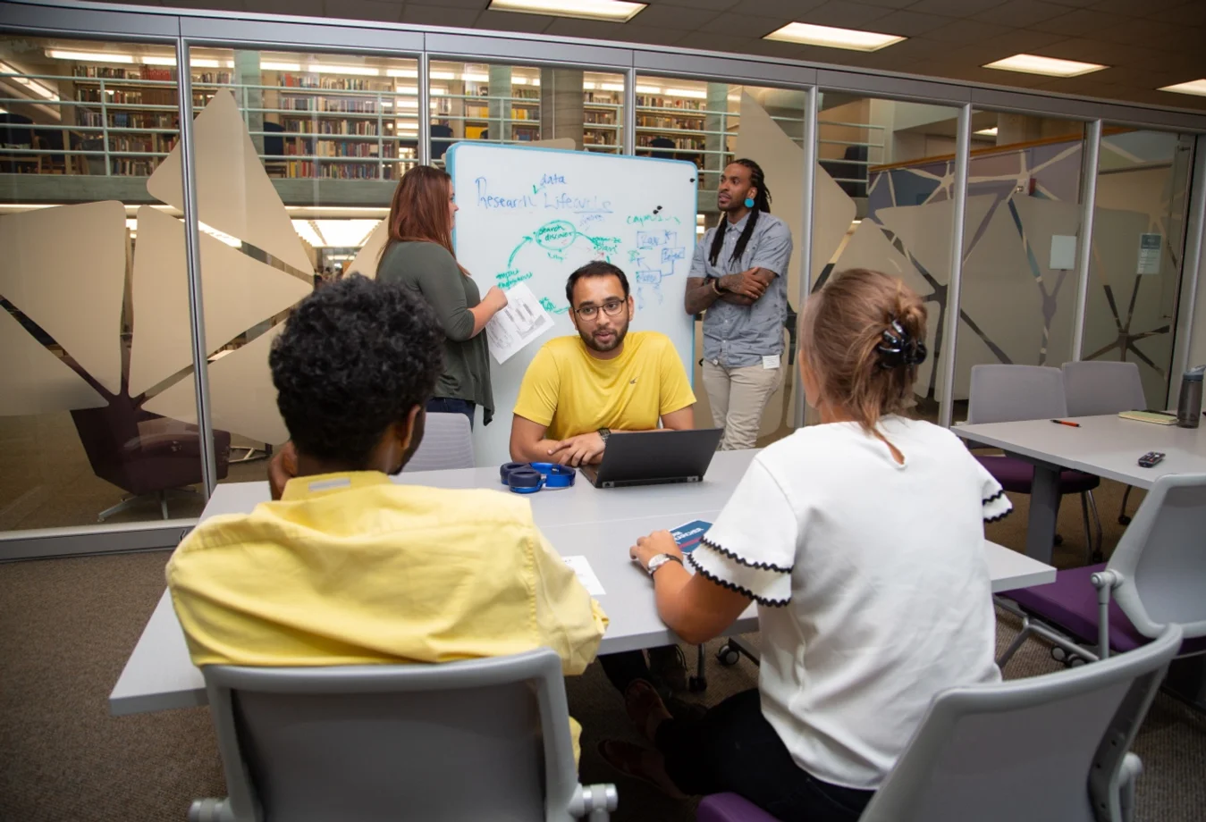 Two students standing by a white board while three students site at a table.