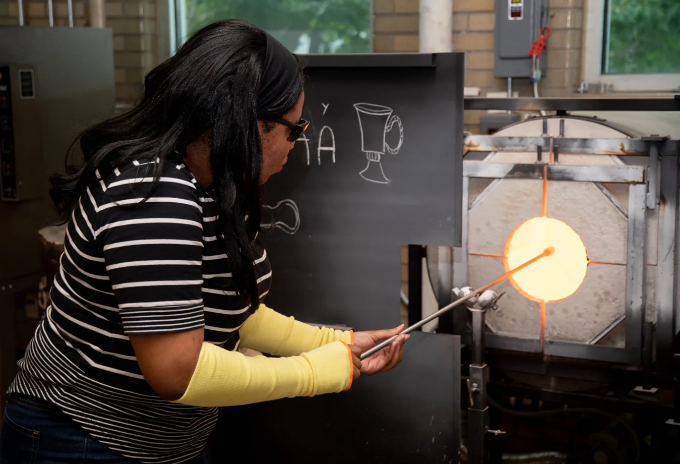 A female student is engaging in the meticulous art of glassblowing, holding a pipe near a furnace known as a 