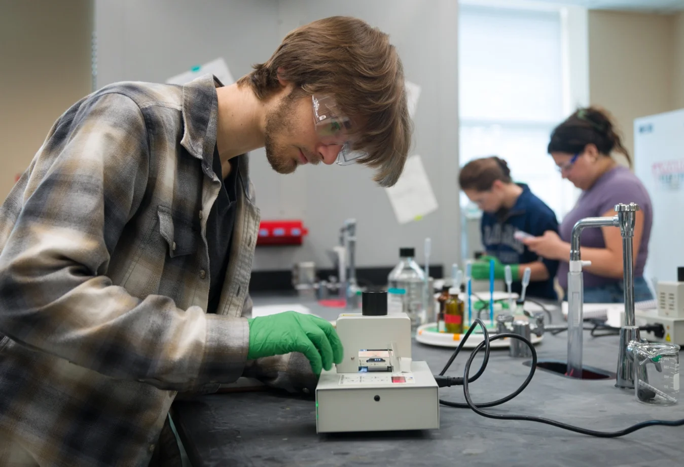 A student wearing safety glasses and green gloves while operating a scientific device in a laboratory setting