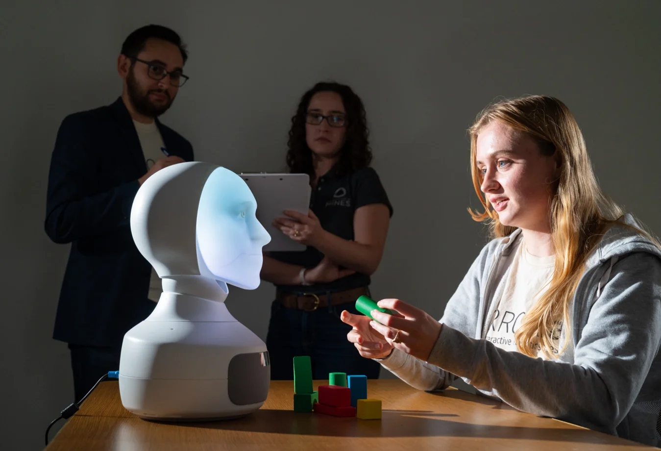A student working with robot with a digital imprint face showcase different shaped blocks as two research students observe.