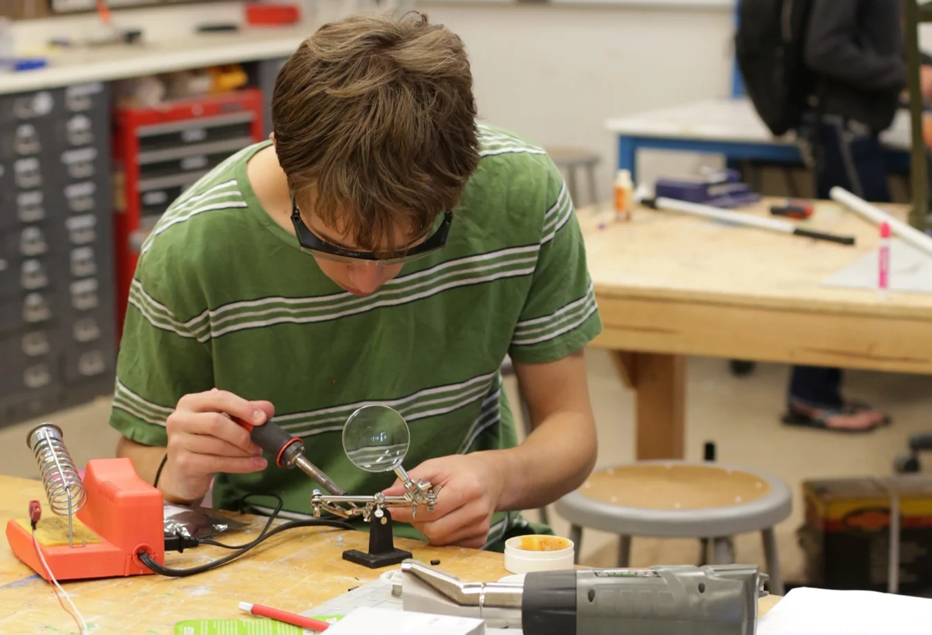 A male Mines student wearing protective glasses as he is working to solder  a circuit board