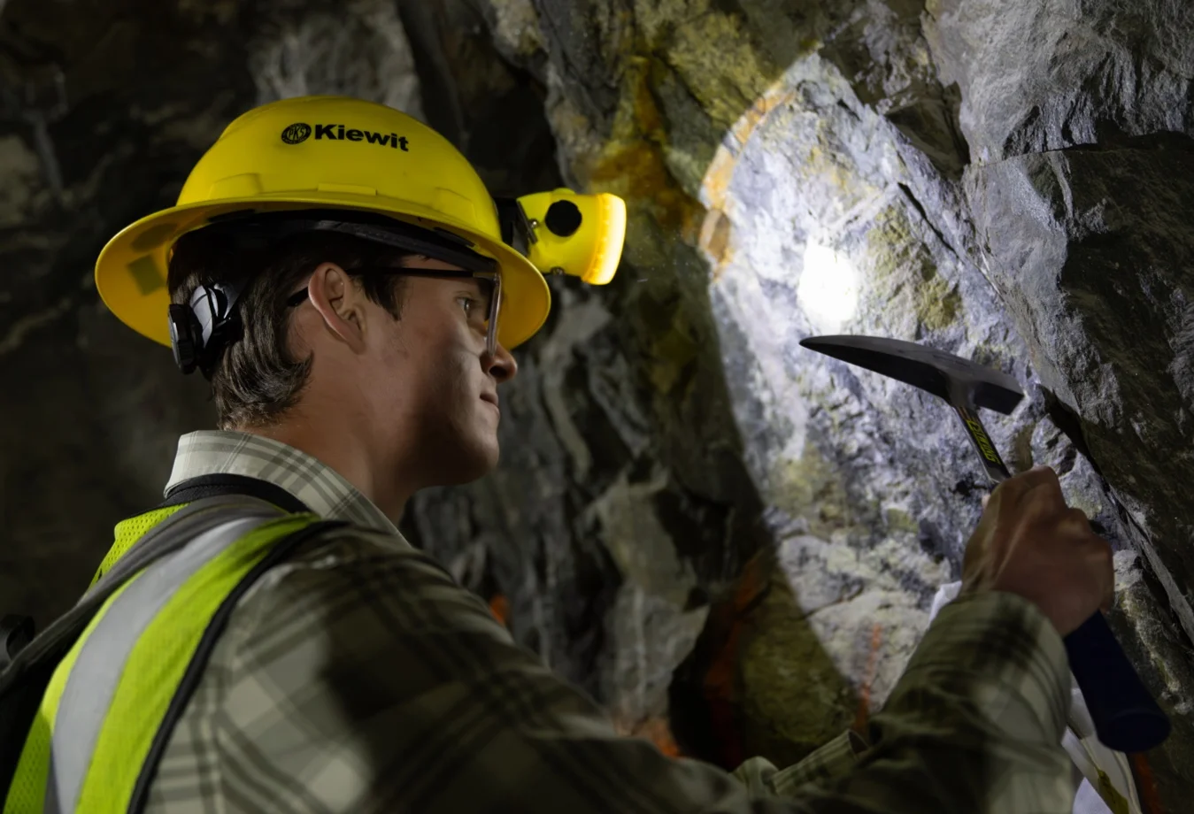 A Mines male student wearing a Kiewit branded yellow hard hat and a high-visibility safety vest, using a pickaxe to conduct field testing in a underground tunnel.