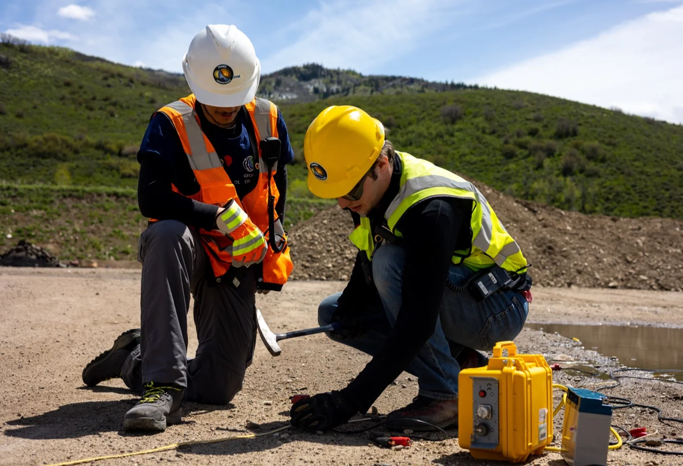 Two male Mines students wearing safety gear conducting geological testing in a field.