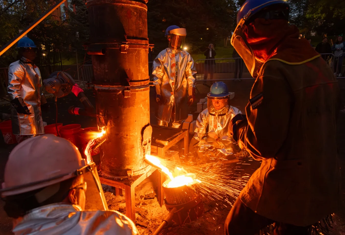 depicts a community iron pour event, a hands-on art workshop where molten metal is cast into molds