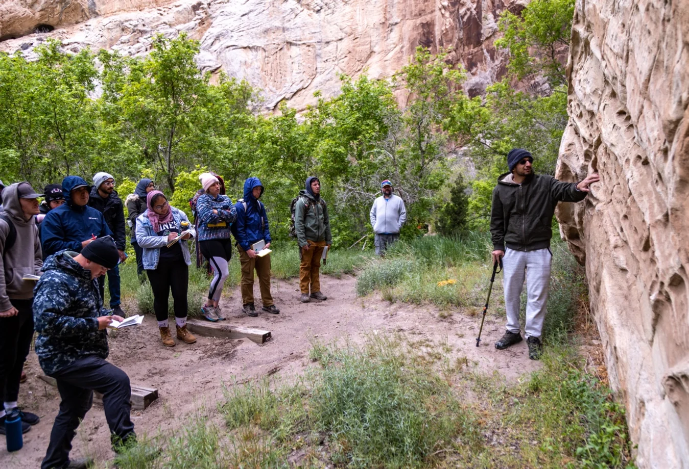 A group of Mines students hiking within a canyon and then listening to their professor explain and examine an canyon rock wall.