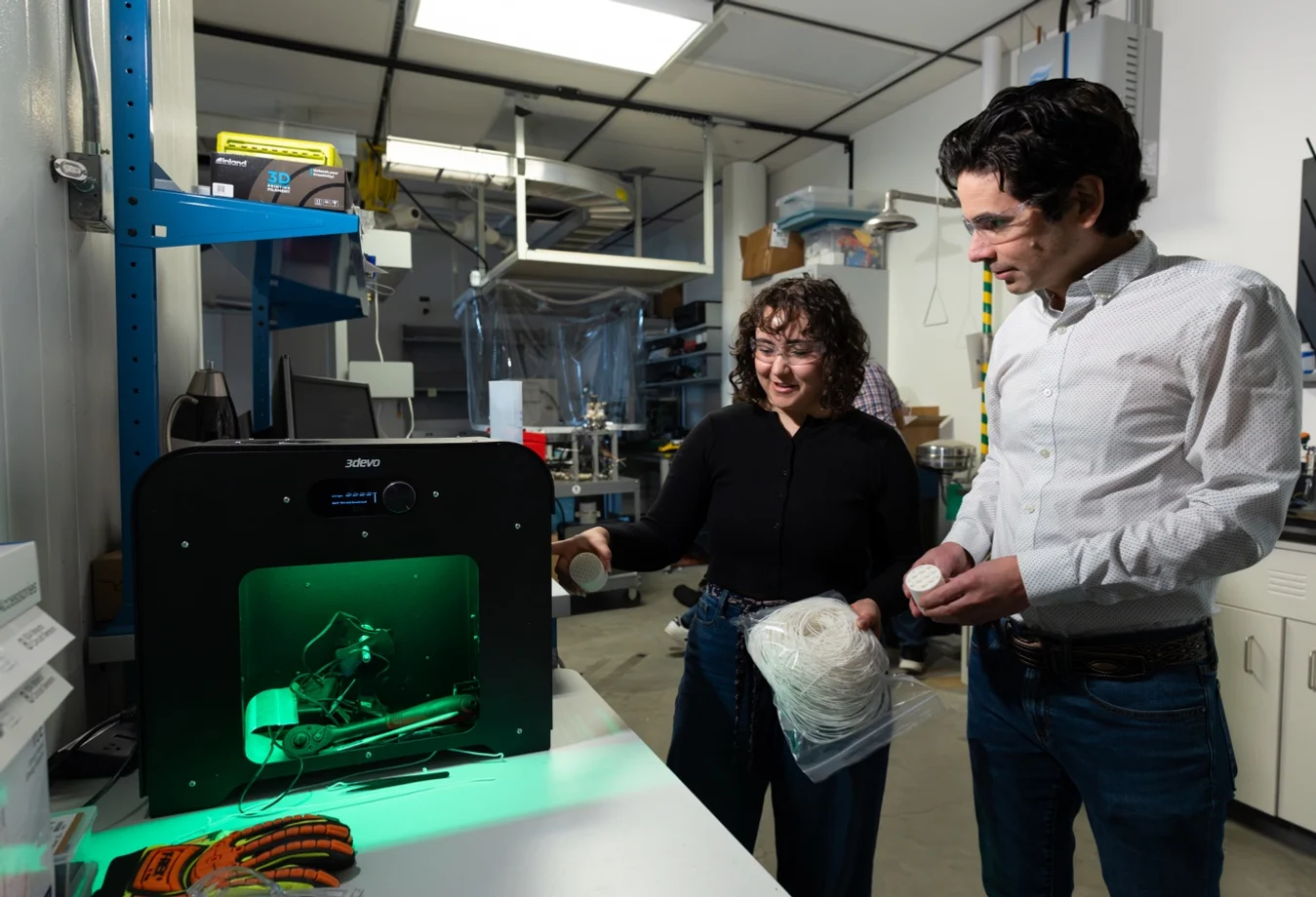 A male and female looking at a 3devo filament maker on a white table, glowing with green light while processing white plastic filament.