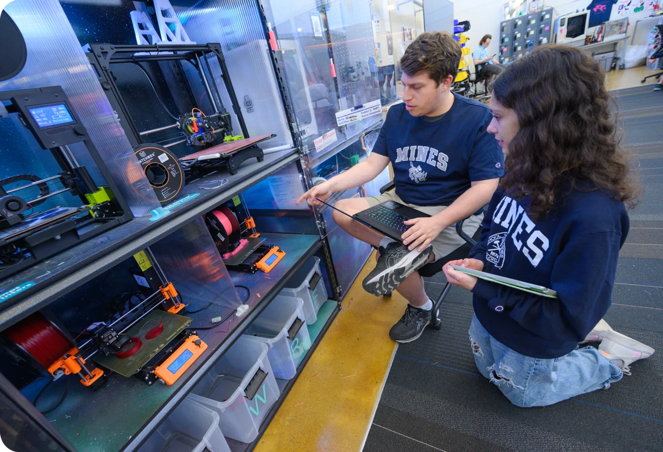 Two Mines students kneeling down in from of 3D printers to confirm the output is correct.