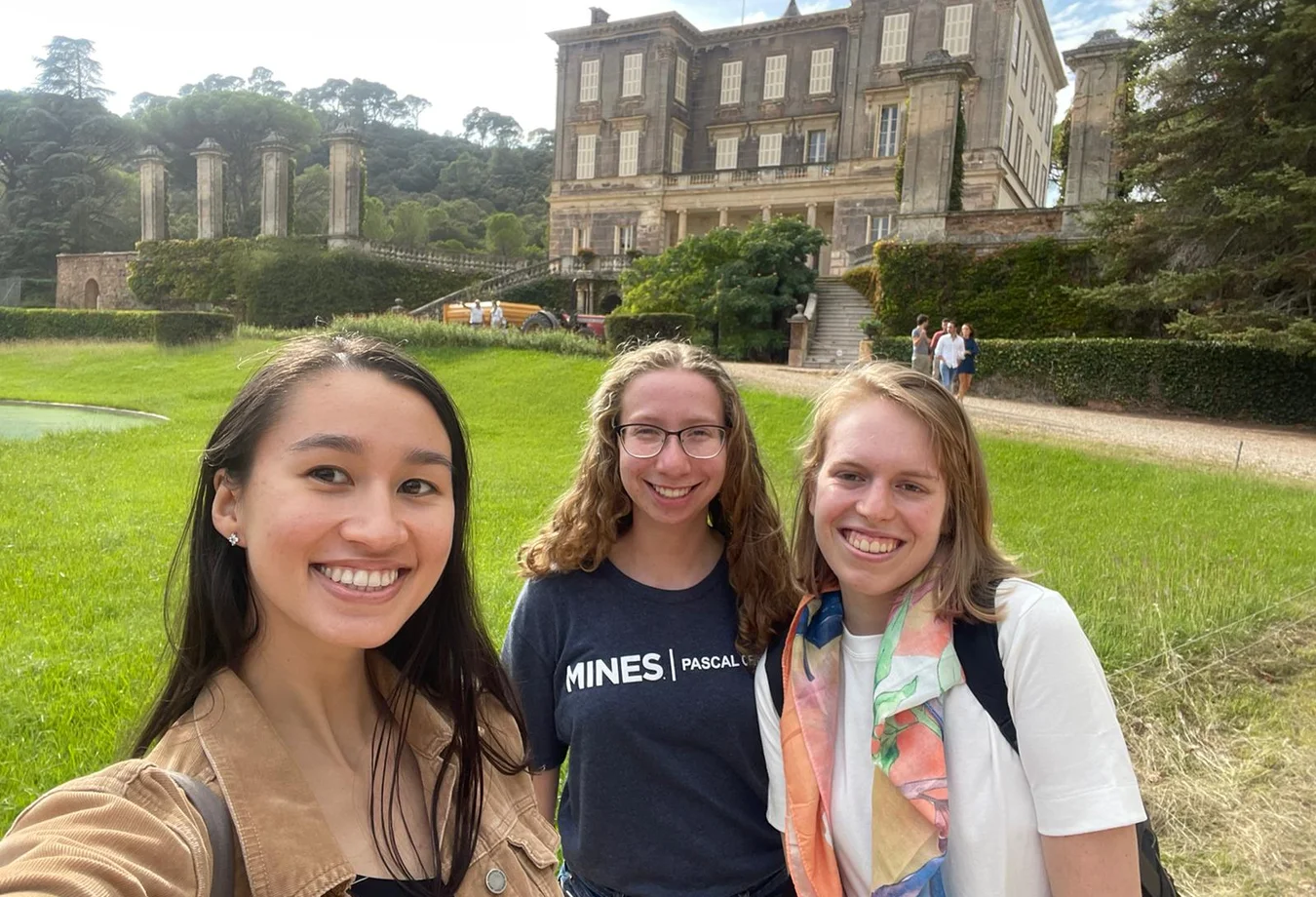 Three women smile for a selfie in front of a large, modern building with glass windows.