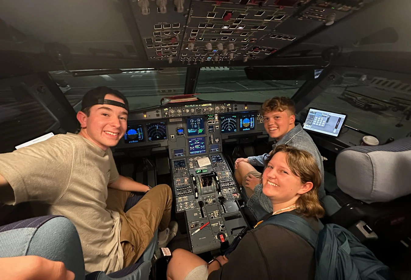 Three young people smiling inside an airplane cockpit surrounded by control panels and screens.