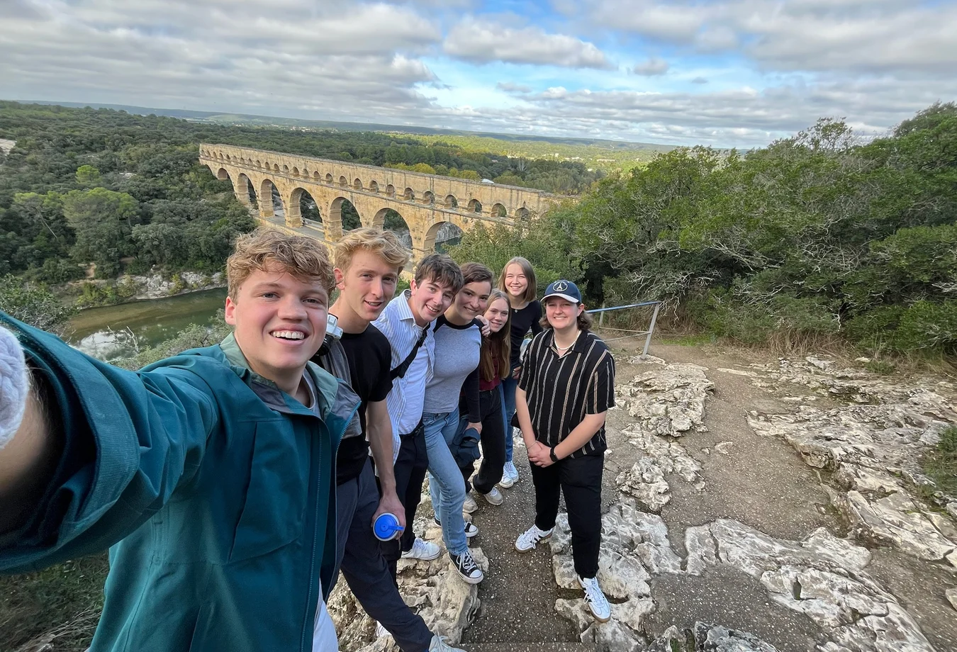Group of seven students taking a selfie on rocky terrain with a historic stone aqueduct and forest in the background under a cloudy sky.