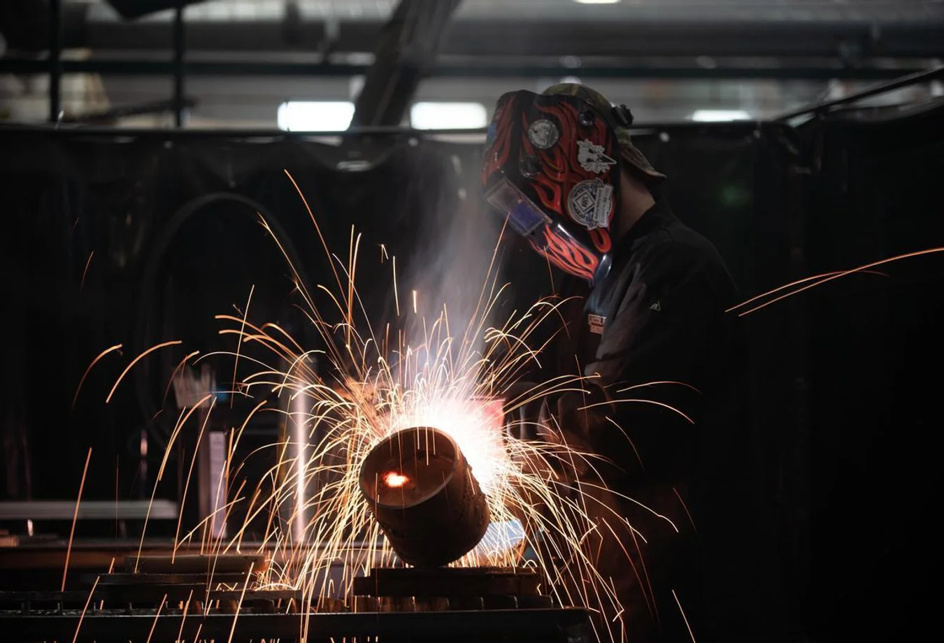 Person welding metal with a protective helmet, creating bright sparks in a dimly lit industrial workshop.