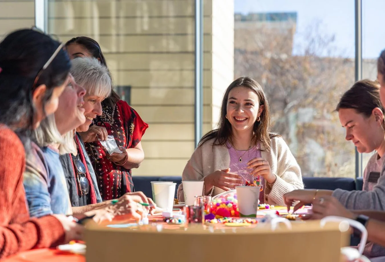 Group of women happily crafting with beads and art supplies around a table in a sunlit room.