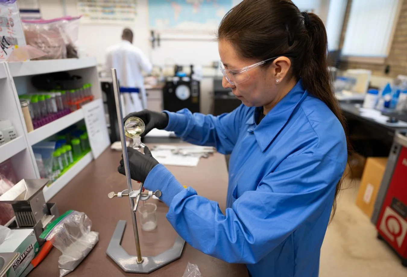 Scientist in blue lab coat and safety glasses pouring liquid into a funnel in a laboratory setting.