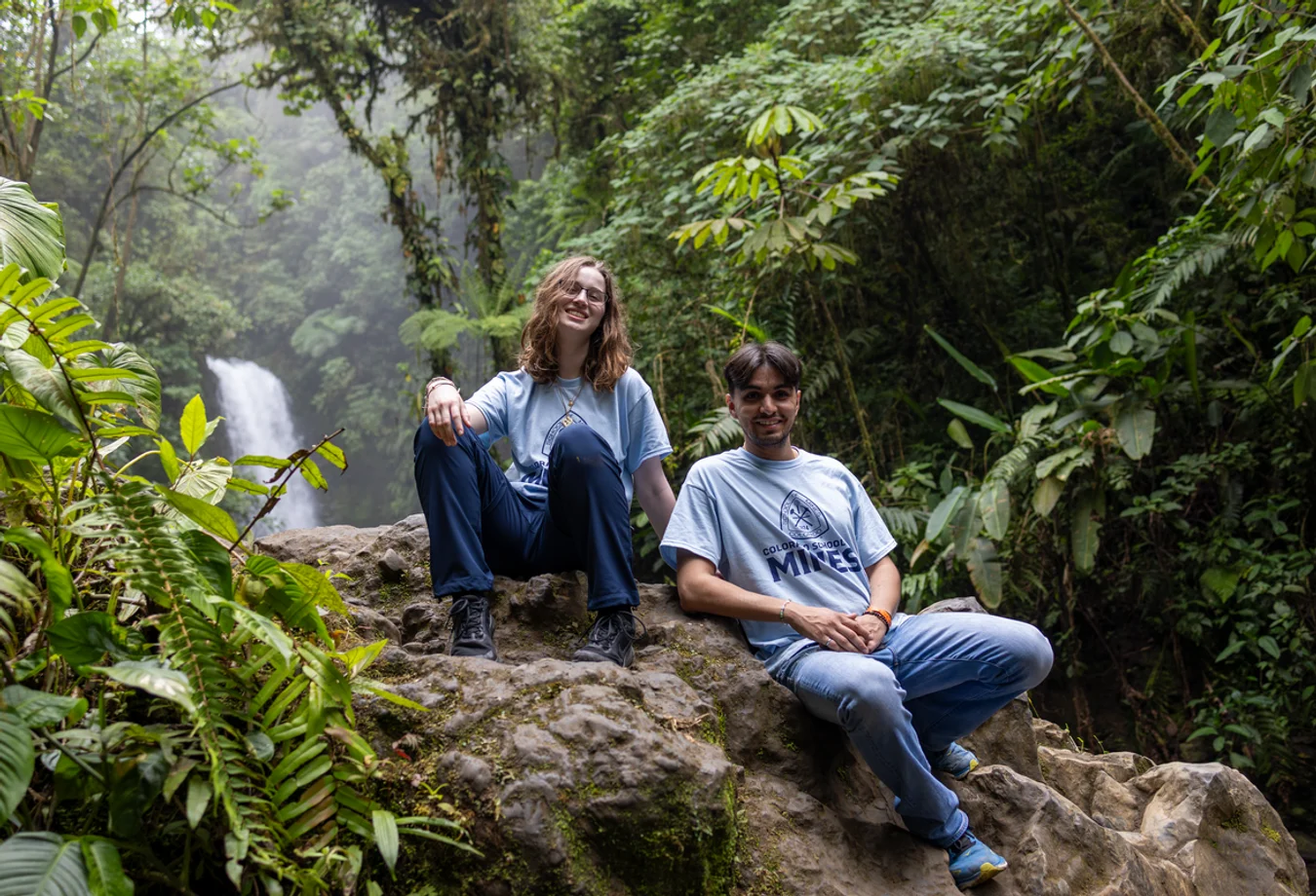 Two Mines students sitting on rocks in a lush, green forest with a waterfall in the background.