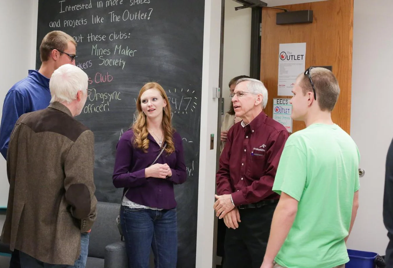 Group of five people having a discussion near a chalkboard with club information and a door labeled 