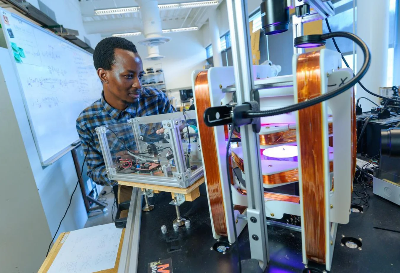 Mines student adjusting advanced lab equipment with copper coils in a bright laboratory setting, with notes on a whiteboard in the background