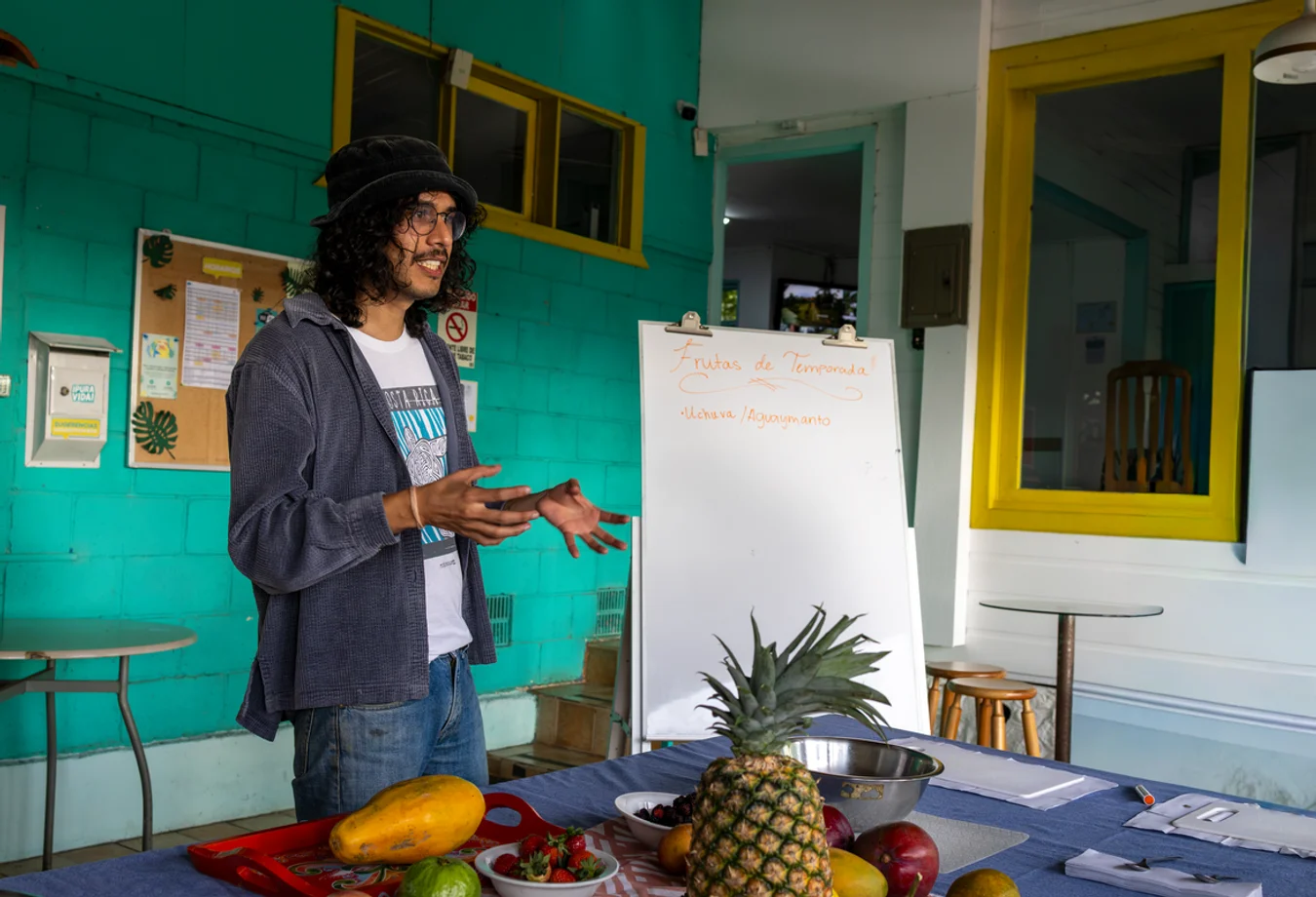 Man with curly hair and glasses giving a presentation about seasonal fruits in a colorful room with tropical fruit on the table.