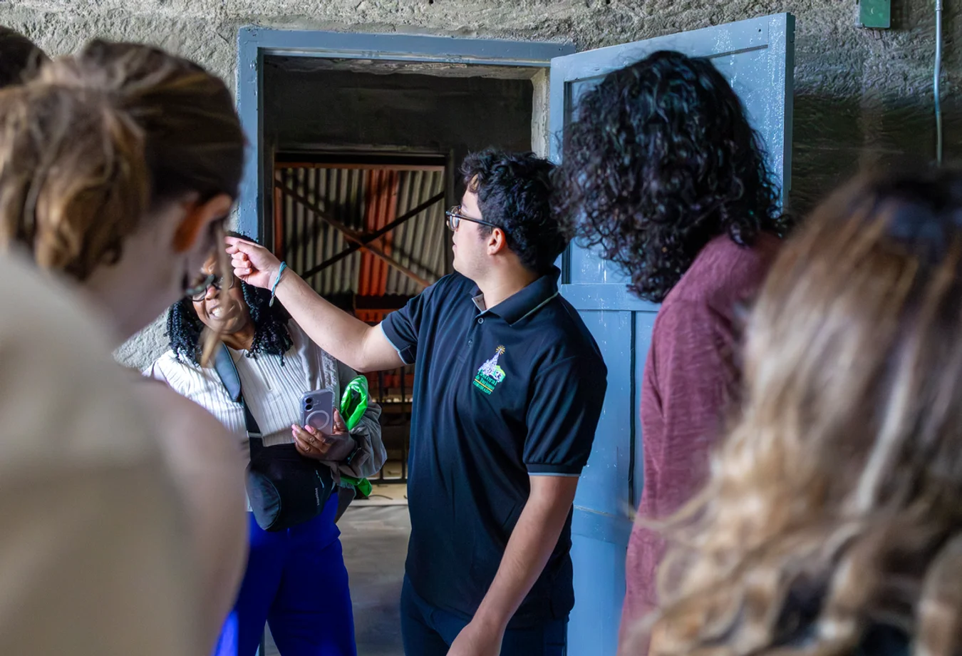 A man in a black polo shirt is pointing while talking to a group of people inside a stone building with a blue door open.