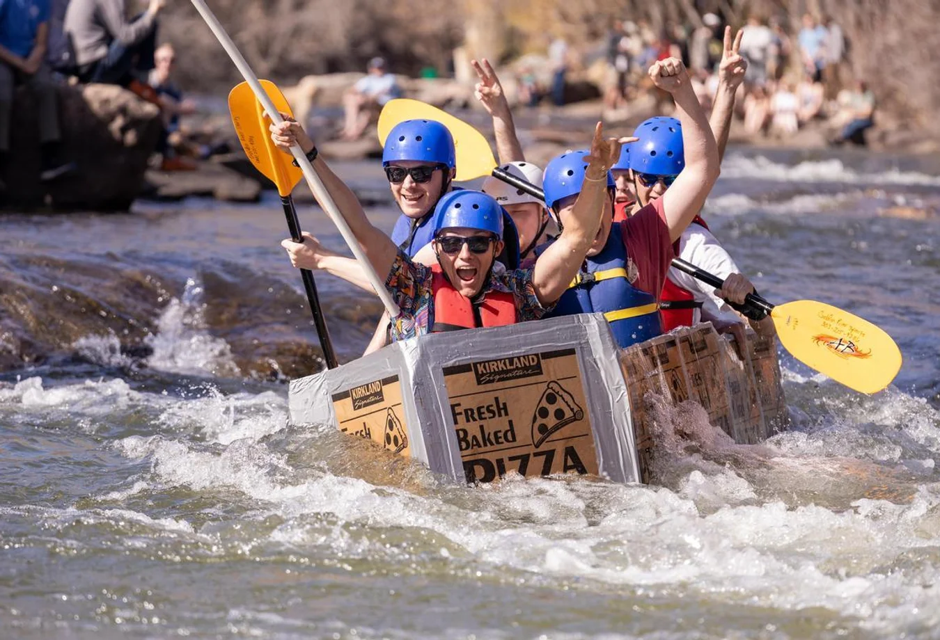 A group of six Mines students wearing helmets and life jackets paddling a cardboard boat made from pizza boxes in a river