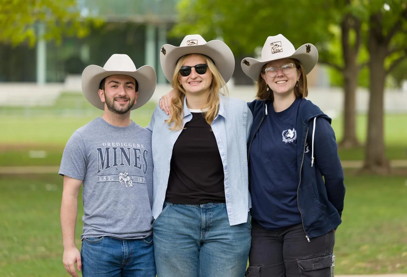 Three people wearing white cowboy hats smiling outdoors with trees and grass in the background.
