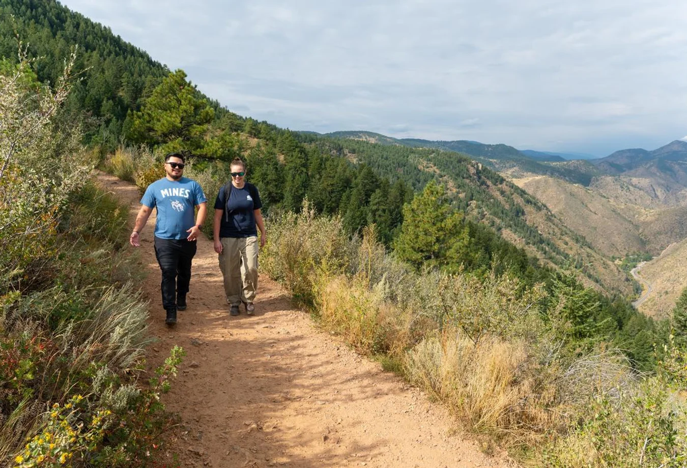 Two people hiking on a dirt trail surrounded by trees and mountains under a partly cloudy sky.