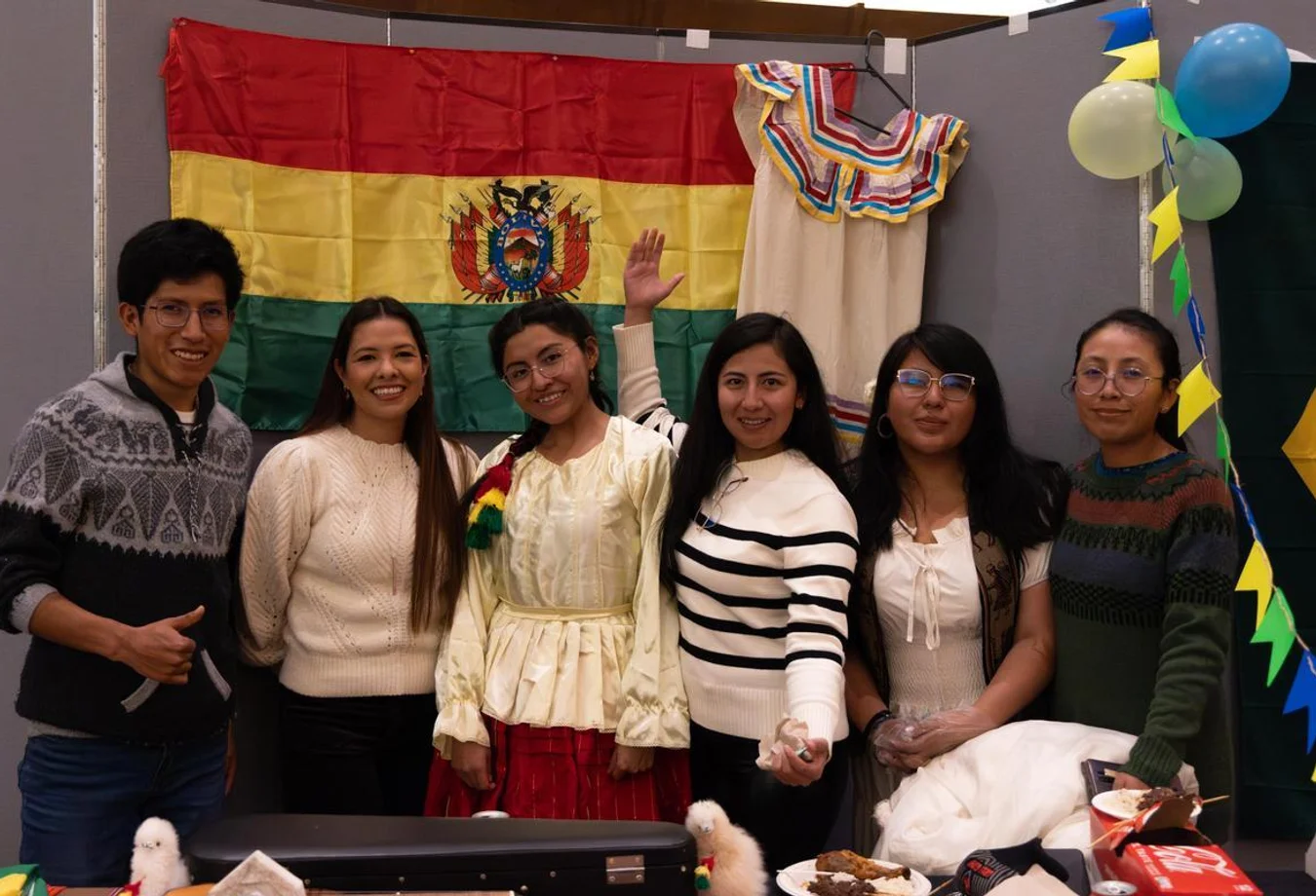 Six students smiling in front of a Bolivian flag with traditional clothing and festive decorations in the background.