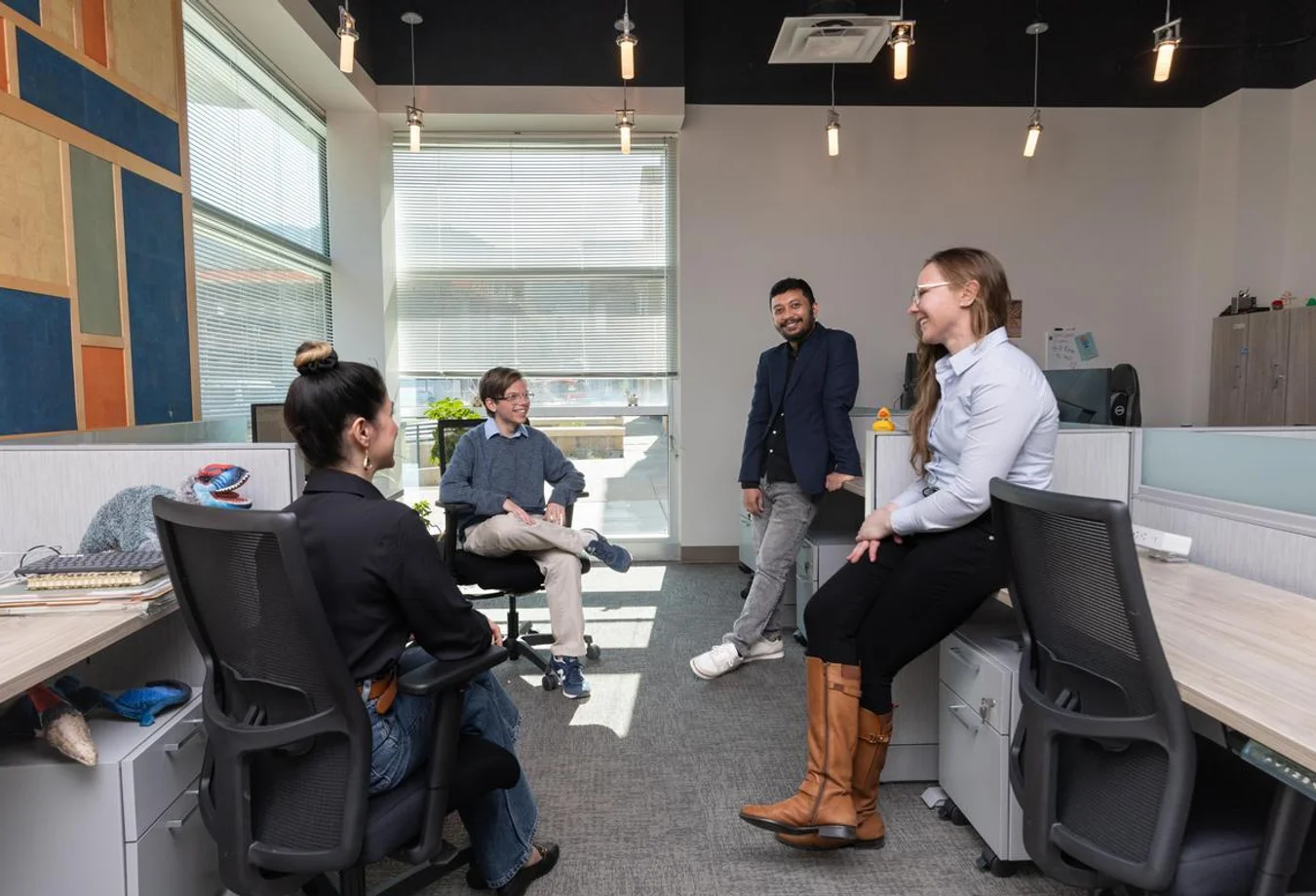 Four coworkers casually chatting and smiling in a modern office space with desks, chairs, and natural light from large windows.