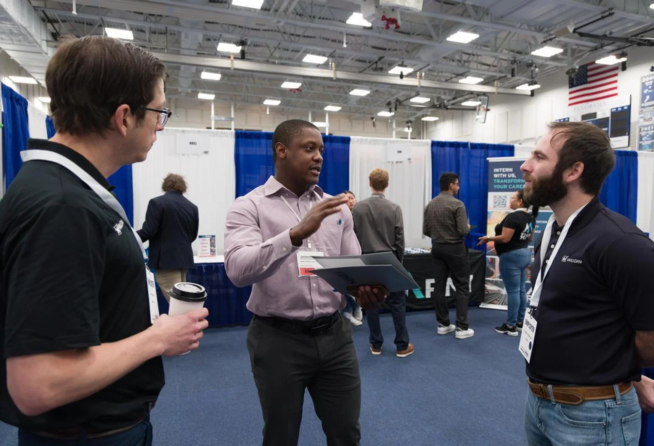 Three men wearing name badges engage in a conversation at Mines Career Days in an indoor venue with booths and an American flag overhead.