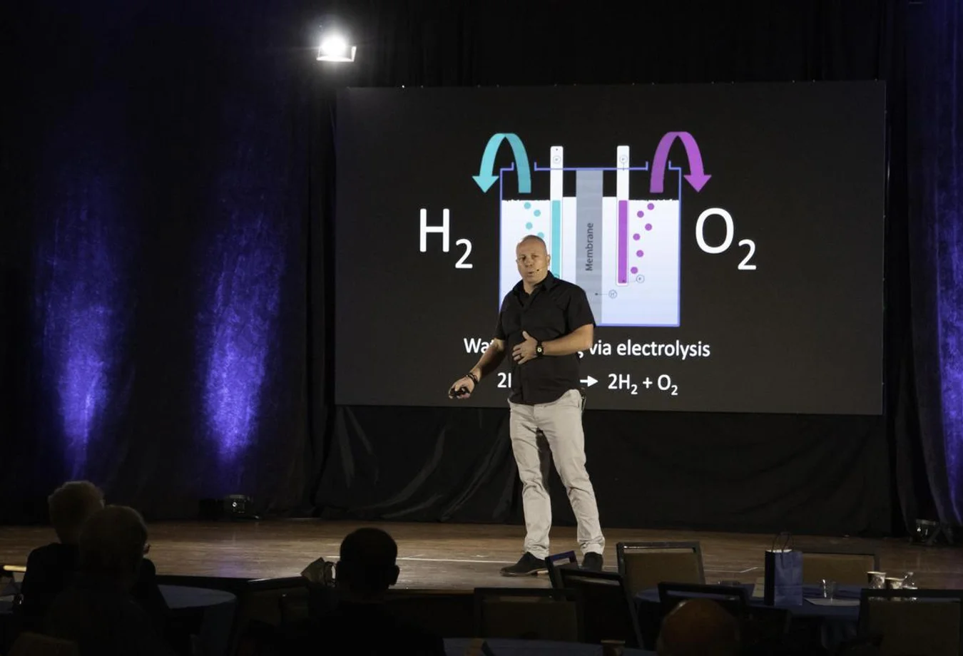 Presenter explains water electrolysis process with H2 and O2 gases shown on screen in a dark conference room with blue lighting.
